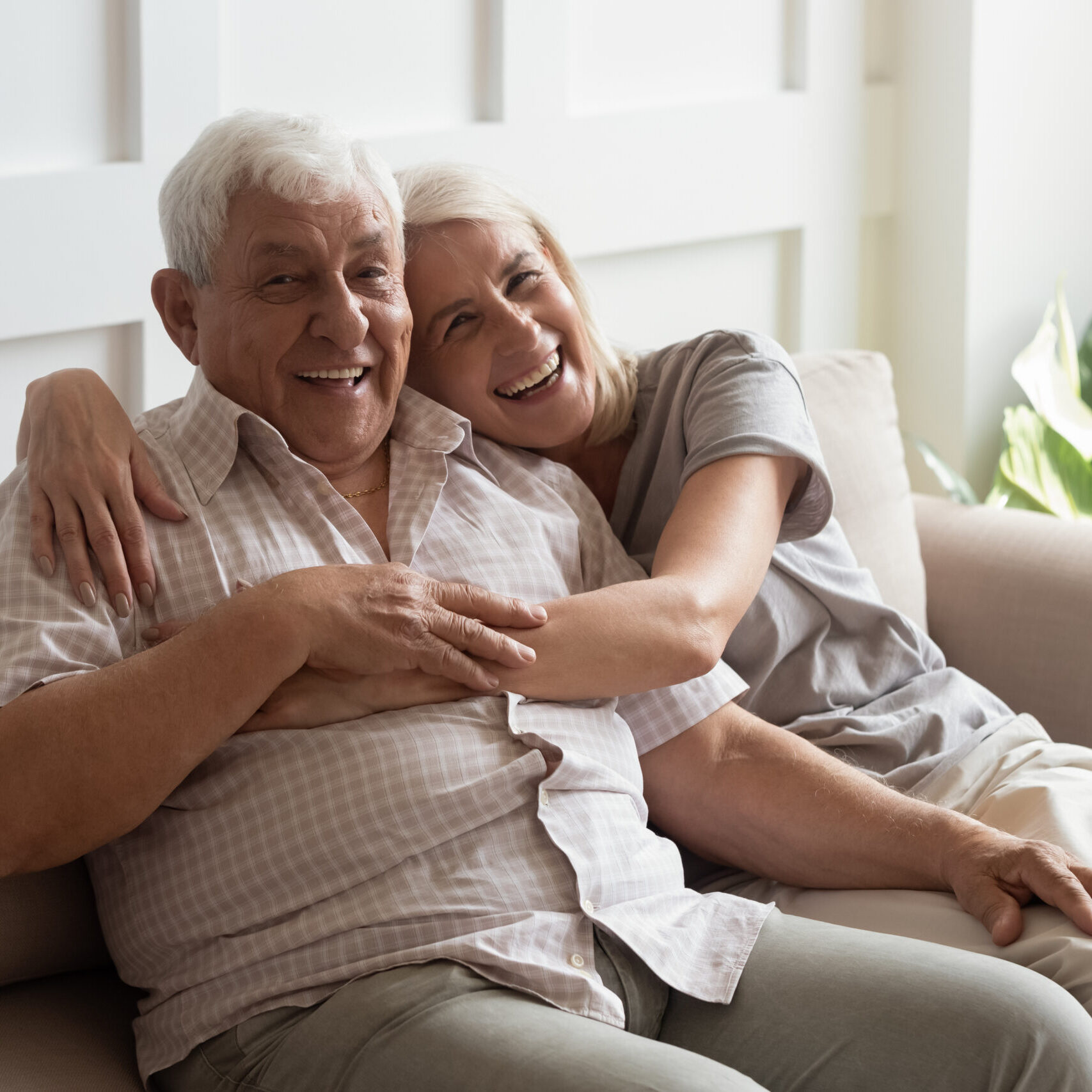 Happy mature couple resting on couch looking at camera Mature healthy couple sitting on couch laughing looks at camera, 80s grey-haired husband 60s wife spend time together feels happy having happy marriage still in love despite the age difference concept